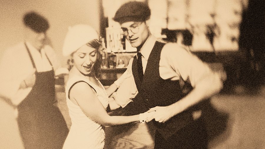 Pareja baila charlestón en un bar de los años 20.  Ella luce vestido claro y gorro; él, camisa, chaleco oscuro y boina. Ambiente festivo.