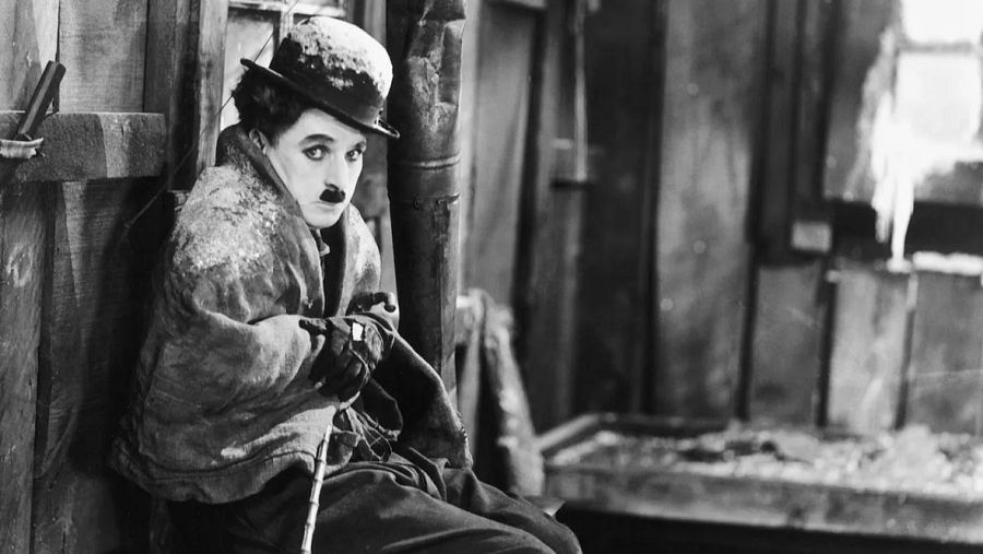 Charles Chaplin, con sombrero y abrigo, sentado en una cabaña nevada.  Expresión triste.  Imagen de "La quimera del oro".