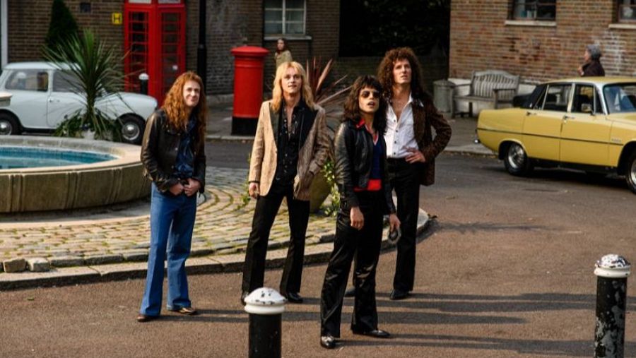 Cuatro actores caracterizados como la banda Queen en una calle con una fuente, cabina telefónica roja y un coche clásico amarillo. Vestuario y ambientación de los años 70.