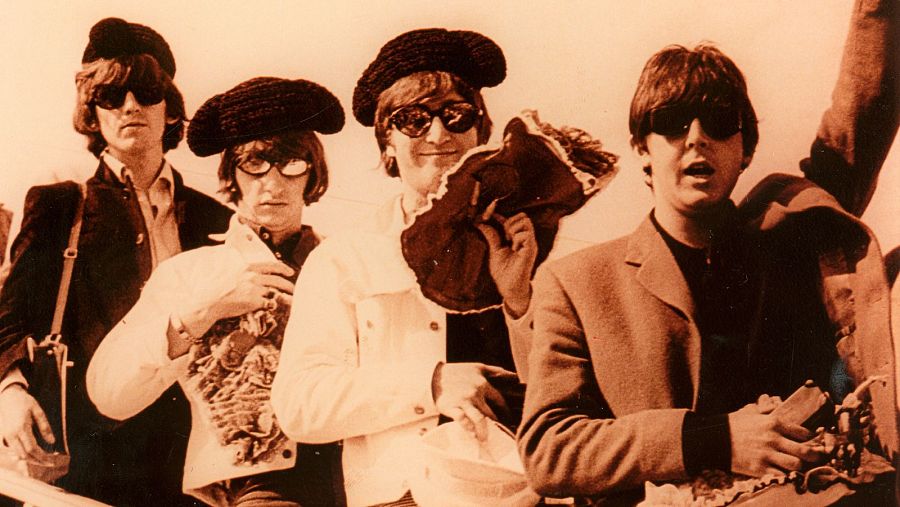 Los Beatles, con boinas y gafas de sol, llegan a España en 1965.  Imagen sepia mostrando a los cuatro miembros de la banda sosteniendo objetos.