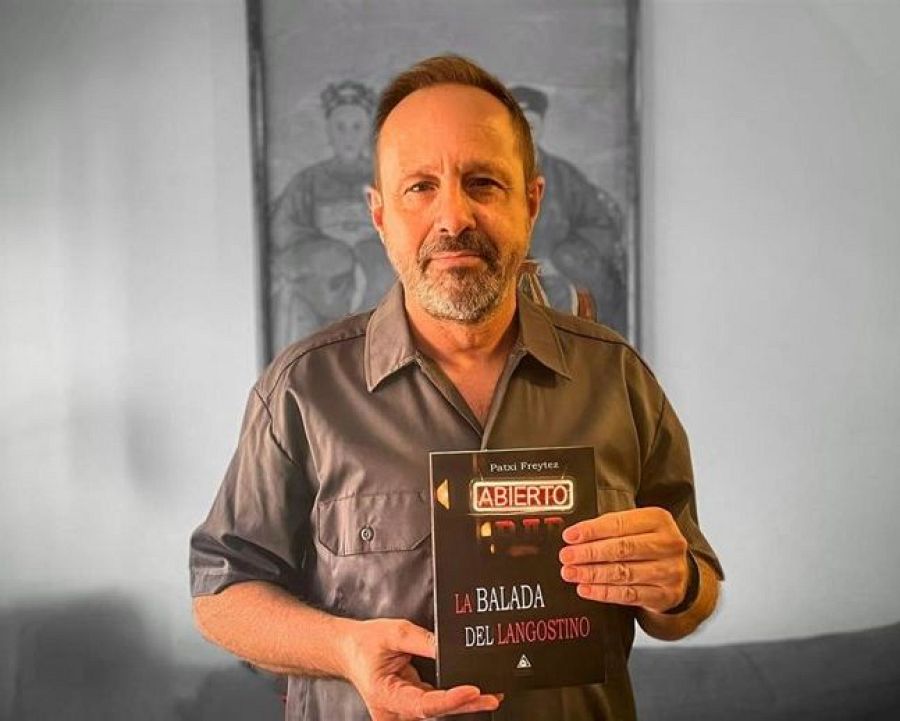Autor con su libro "La balada del langostino", portada con "ABIERTO" en estilo luminoso.  Hombre de mediana edad, camisa gris oscura, cabello canoso.