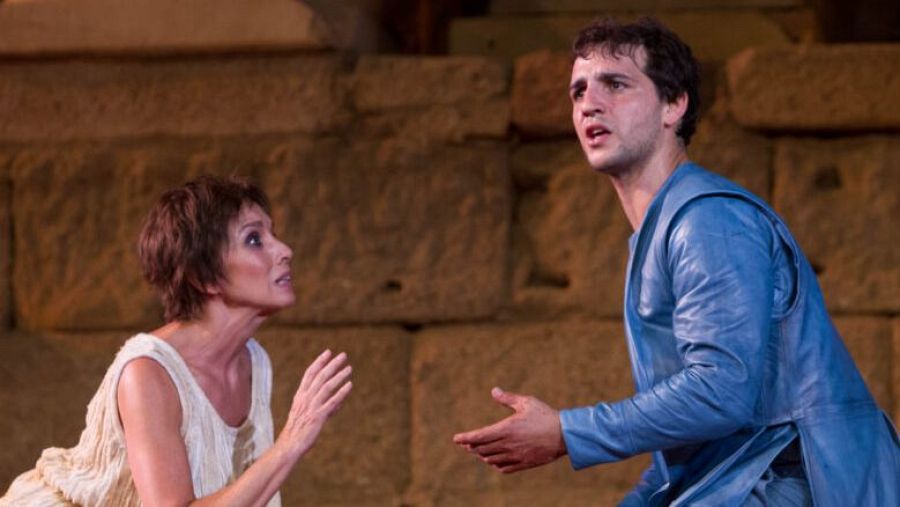 Intensa escena de "Electra" en Mérida 2025: dos actores, ella con túnica clara y él con túnica azul, expresan tensión en un escenario de piedra.