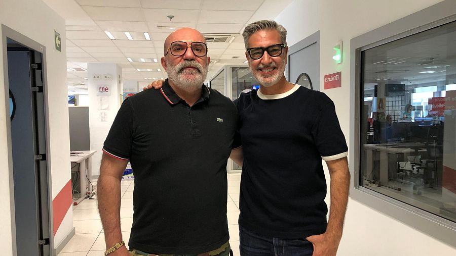 Dos hombres, uno con polo negro y pantalón de camuflaje, el otro con camiseta azul, conversan en un pasillo con el logo de RNE visible.  Se observa el cartel "Estudio 105".