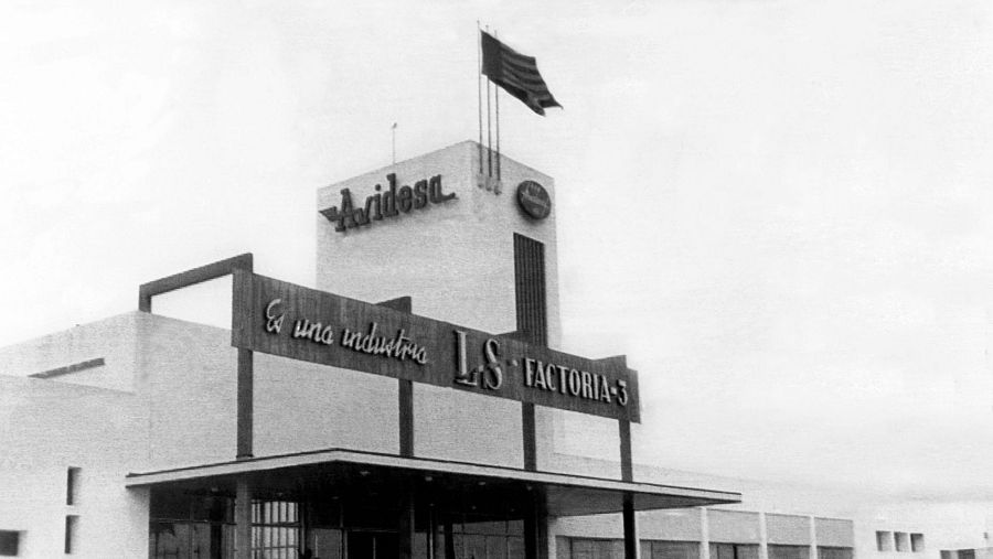 Fachada de fábrica antigua con rótulos, bandera española y las iniciales "LS".  Estilo arquitectónico clásico. Fotografía en blanco y negro.