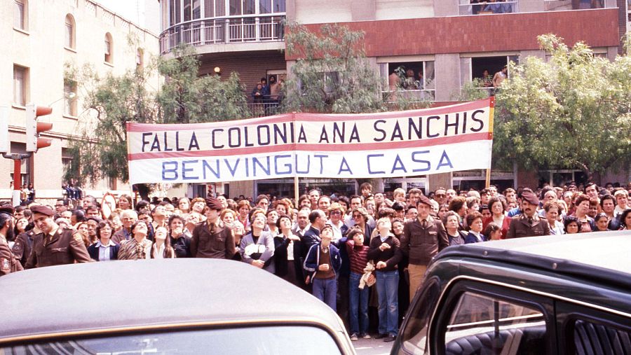 Multitud en Alzira con pancarta "Falla Colonia Ana Sanchis Bienvenido a casa", esperando la llegada de alguien.