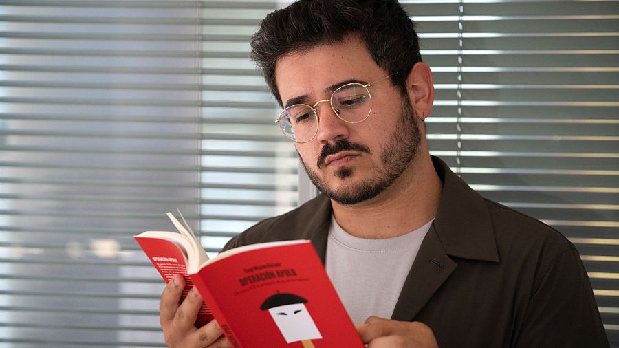 Hombre con gafas leyendo un libro rojo titulado "Operación Apolo", con ilustración de una persona con boina.  Imagen tomada frente a una ventana con persianas.