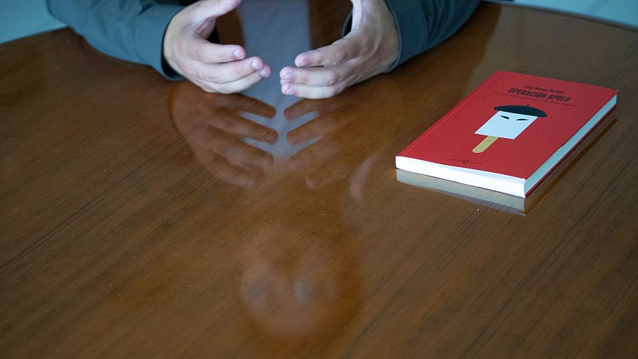 Entrevista a Sergi Moyano en Valencia.  Sus manos descansan sobre una mesa de madera pulida, junto a un libro rojo titulado "Operación Apolo".