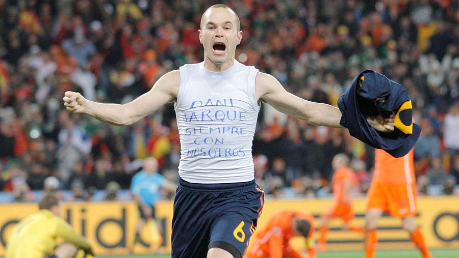 Iniesta celebra con una camiseta que reza "DANI JARQUE SIEMPRE CON NOSOTROS", puño cerrado y una camiseta azul en la mano. Estadio repleto.