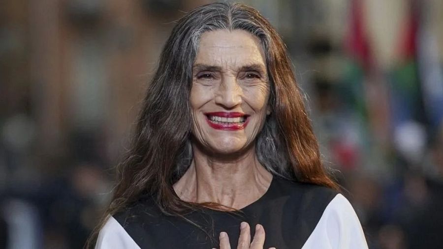 Actriz con cabello castaño y canas, sonriendo, viste un atuendo blanco y negro en una alfombra azul. Fondo desenfocado.