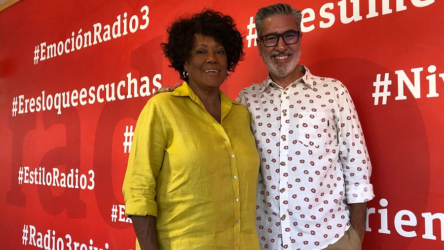 Dos personas, un hombre con camisa blanca y gafas y una mujer con camisa amarilla, posan ante un cartel rojo con hashtags de Radio 3 en RNE Madrid.