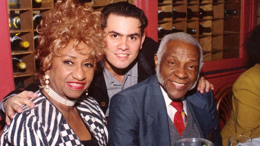 Celia Cruz y dos hombres sonríen a la cámara en lo que parece un restaurante.  Ella viste una chaqueta a rayas y un collar de perlas; los hombres llevan chaquetas y camisas.
