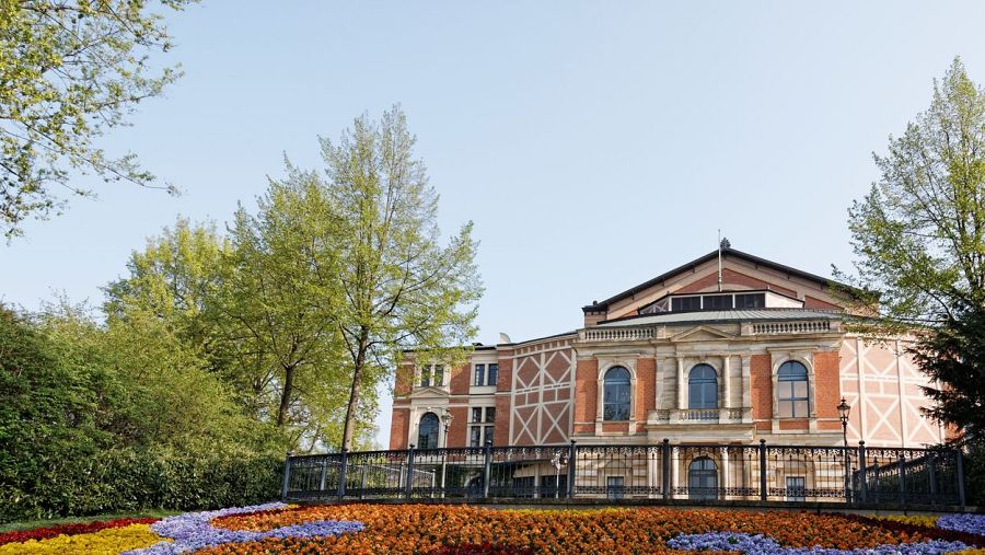 Fachada de teatro de ladrillo rojizo y piedra clara, con columnas, ventanas arqueadas y un jardín de flores multicolores en primer plano.  Cielo azul despejado y árboles verdes al fondo.