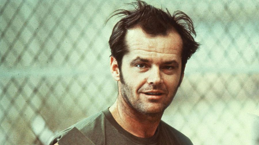 Jack Nicholson, como Randle McMurphy en "Alguien voló sobre el nido del cuco", con una expresión pícara y camiseta verde oliva. Fondo borroso con textura metálica.