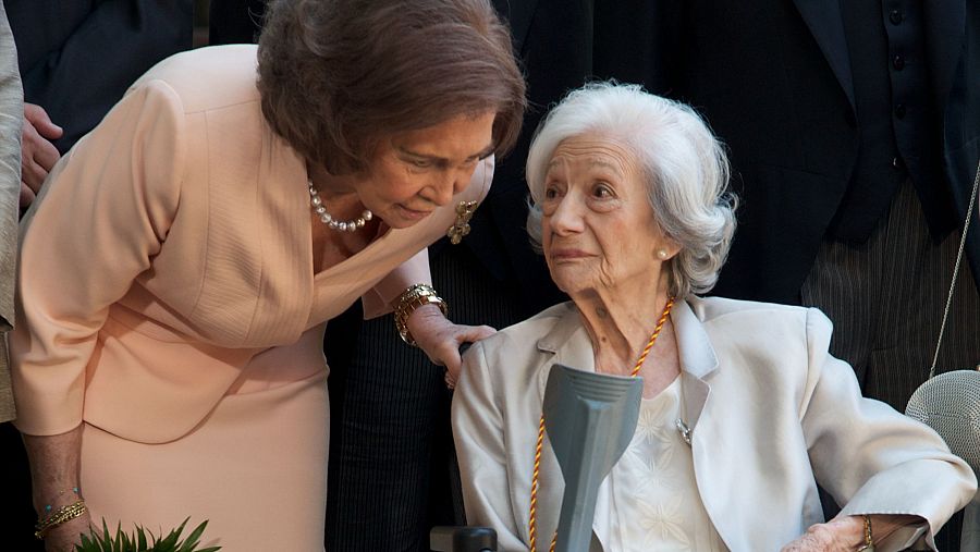 Fotografía de la Reina Sofía y Ana María Matute en la entrega del Premio Cervantes 2010. La Reina, con traje melocotón y collar de perlas, conversa con Matute, sentada.