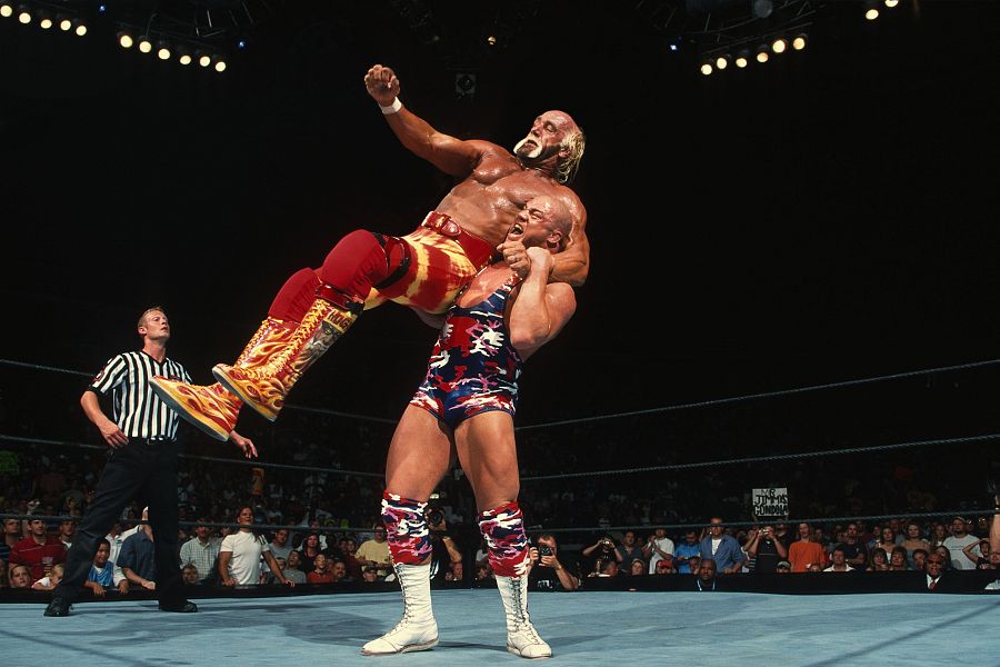 Combate de lucha libre: Angle, en malla de camuflaje, alza a Hogan, quien viste botas con llamas y mallas rojas y amarillas. El árbitro observa la escena.