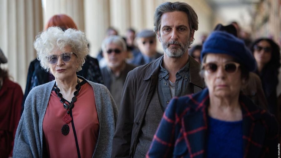 Escena de "Un mundo normal" con Magüi Mira (chaqueta gris, gafas) y Ernesto Alterio (chaqueta marrón) caminando entre gente en un lugar con columnas.
