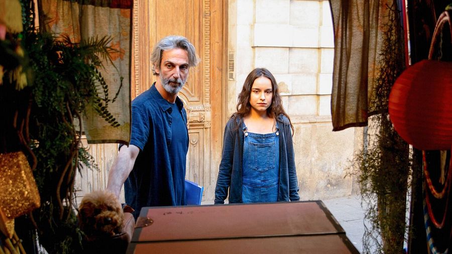 Ernesto Alterio y Gala Amyach en una escena de "Un mundo normal". Él, con barba y camiseta azul, ofrece un peluche a un perro. Ella, con peto vaquero y chaqueta gris, observa con seriedad.