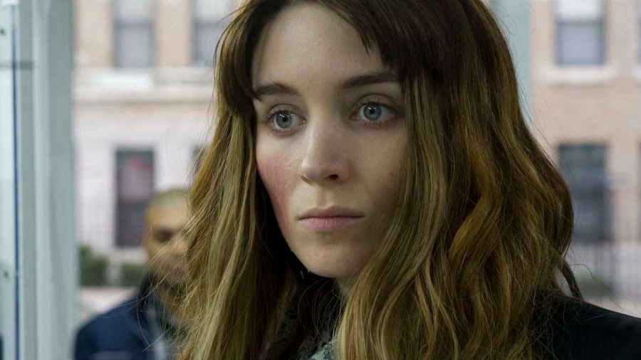 Primer plano de Rooney Mara con expresión seria, pelo largo castaño claro y prenda oscura. Fondo desenfocado con un edificio y otra persona.