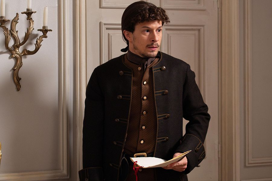 Hombre de época con chaqueta oscura y chaleco marrón, sostiene un documento en un interior elegante con candelabro dorado.  Ambientación que sugiere un momento clave.