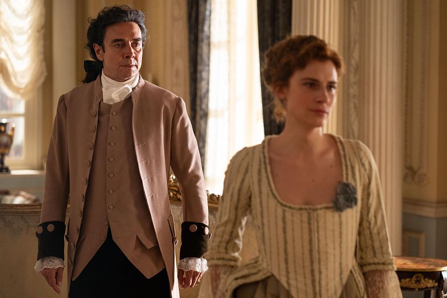 Interior elegante: hombre con levita y mujer con vestido de época se miran con expresiones serias y neutras.  Posible escena de "Valle Salvaje".