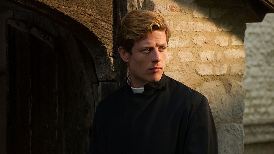 Imagen de medio cuerpo de James Norton interpretando al vicario Sidney Chambers en 'Grantchester'. Viste sotana negra, apoyado en pared de piedra y madera, con expresión seria.