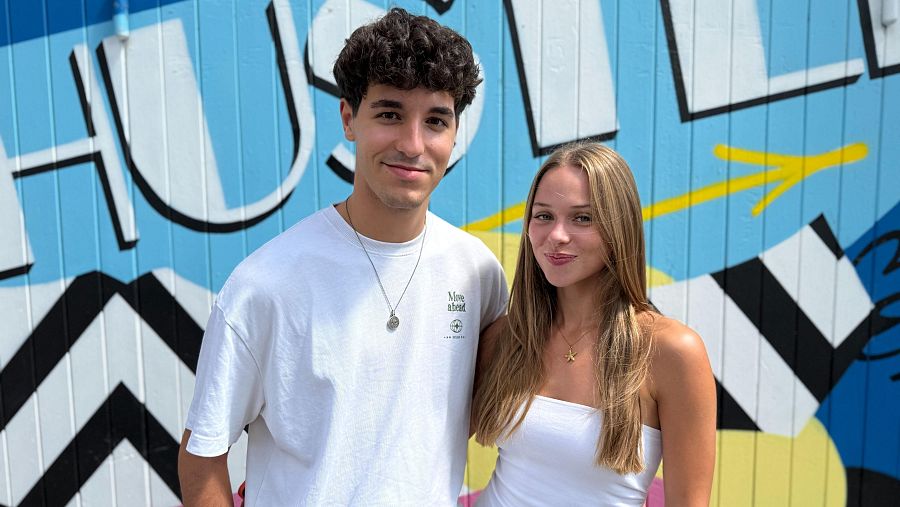 Jóvenes posando ante mural geométrico con la palabra "HUSTLE". Él, pelo rizado y collar circular; ella, pelo rubio y collar con colgante de estrella de mar. Ambos visten ropa clara.