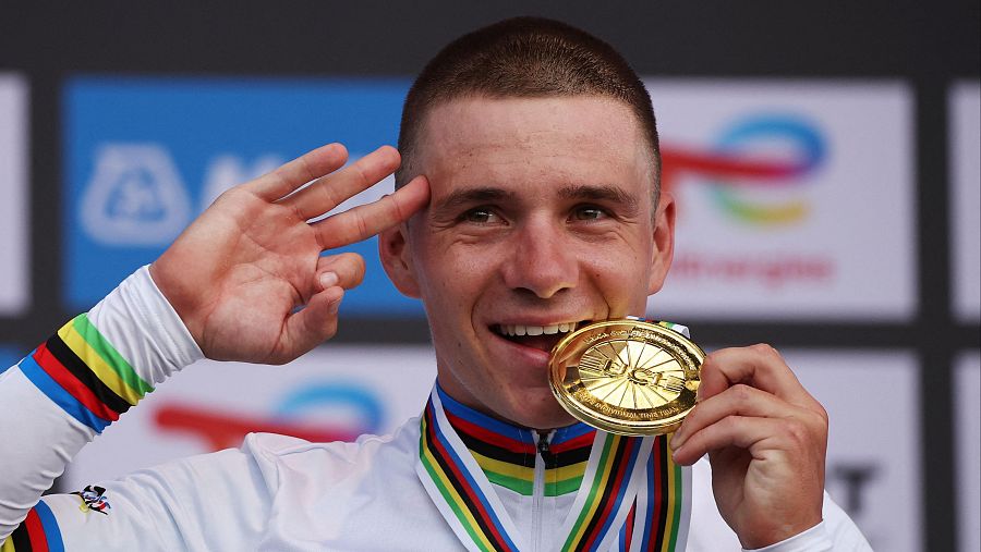 Ciclista con maillot blanco y arcoíris, mordiendo medalla de oro y con la mano en la frente, celebrando una victoria.