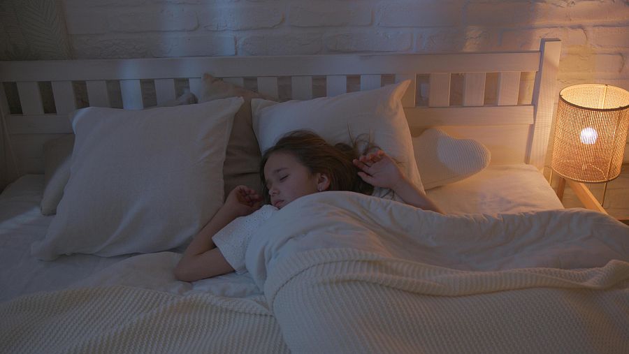 Niña dormida boca arriba en cama blanca con manta, iluminada por una lámpara de mesa cercana. Ambiente sereno y tranquilo.