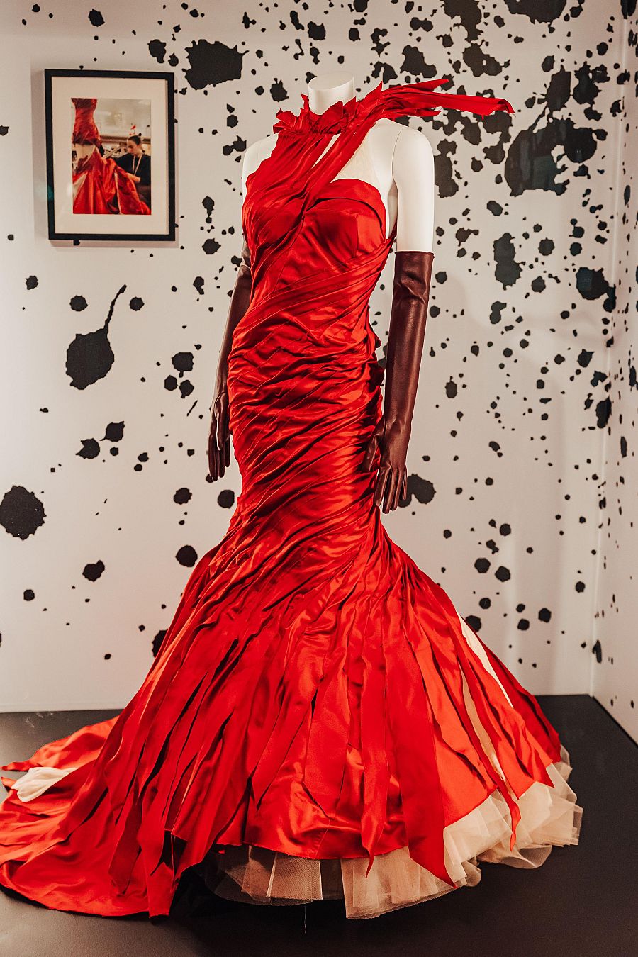 Un maniquí blanco con guantes oscuros exhibe un vestido rojo de gala con una falda larga y detalles de tela. El fondo es una pared blanca con manchas negras y una fotografía enmarcada.