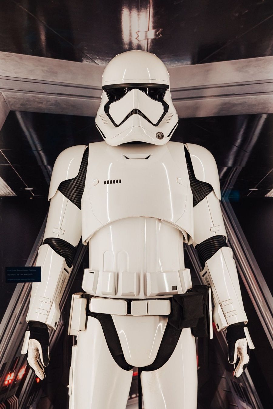 Una armadura blanca de Stormtrooper, con un casco de diseño característico y detalles negros, se muestra en un entorno con líneas horizontales y luz tenue. La armadura cubre todo el cuerpo, incluyendo hombreras y protectores.