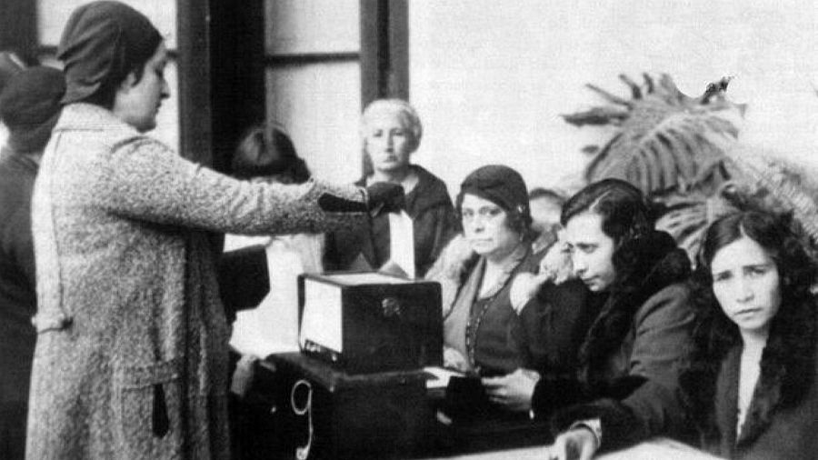 En una escena histórica, una mujer deposita su voto en una urna electoral, mientras otras mujeres esperan su turno, vestidas con ropa de abrigo y sombreros, en un ambiente que sugiere un colegio electoral.