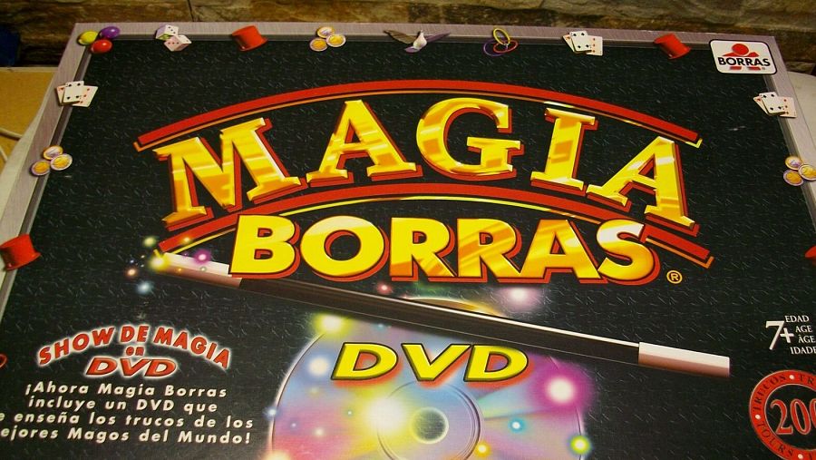 La caja del juego de magia presenta el título "MAGIA BORRAS" en letras grandes, junto con la frase "SHOW DE MAGIA DVD". Se observa una varita mágica y elementos gráficos que sugieren trucos de magia, además de la indicación de edad "7+".