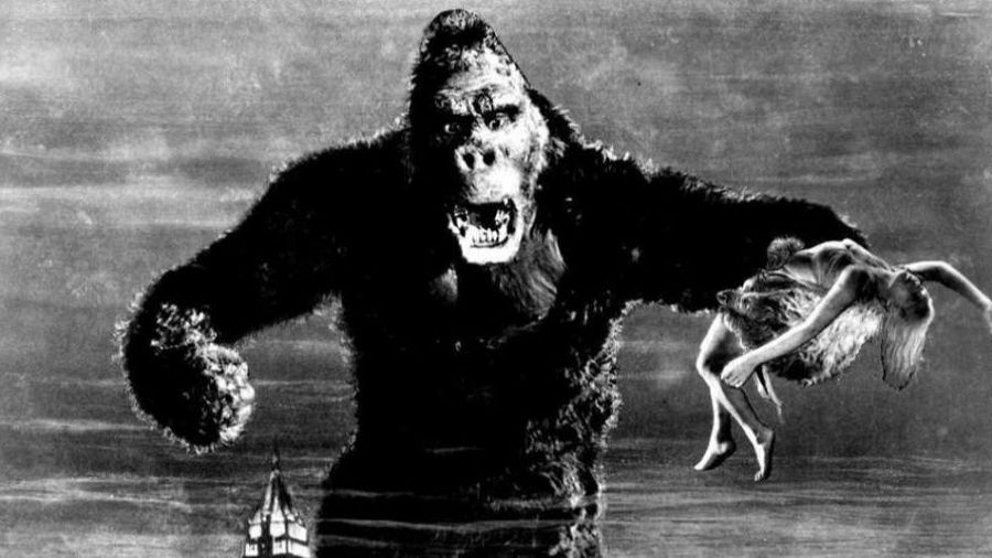 King Kong, un gorila gigante, sostiene a una mujer en su mano, mostrando una expresión amenazante. La imagen en blanco y negro presenta un edificio alto en la parte inferior y un cielo nublado.