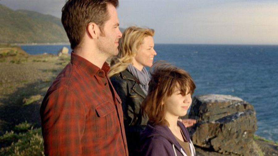 Una familia, compuesta por un hombre con camisa de cuadros, una mujer con chaqueta de cuero y una niña con sudadera azul, disfruta de las vistas al mar desde un acantilado en un día soleado.