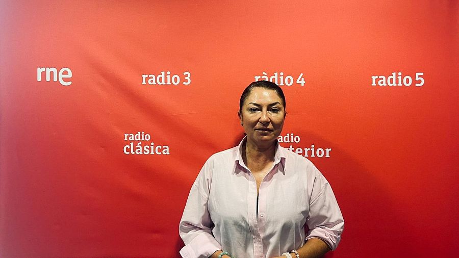 Montserrat Martí, con el pelo recogido y camisa rosa claro, se encuentra frente a un fondo rojo con logotipos de emisoras de radio.