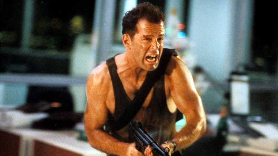 En una escena de acción, Bruce Willis, como John McClane, aparece con el rostro herido y gritando, sosteniendo un arma en el interior de un edificio con vistas a la ciudad.