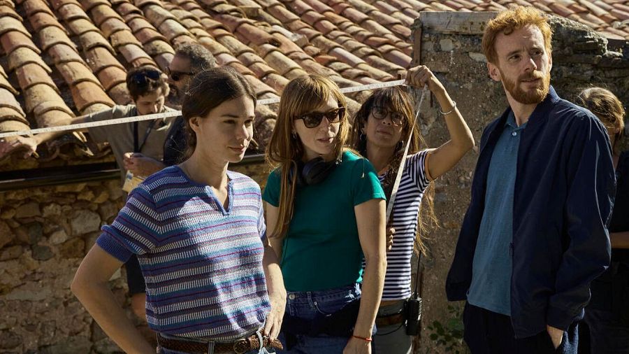 En el set de filmación, un grupo de personas se encuentran en primer plano. Una mujer con camiseta a rayas y otra con gafas de sol y auriculares, junto a una tercera con gafas y camiseta a rayas, sostienen una cinta métrica. Un hombre pelirrojo completa la escena.