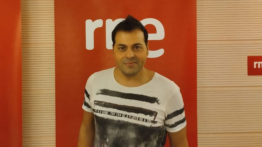 Miguel Herrero, con camiseta estampada y pelo corto, posa frente a un fondo rojo con el logo de una emisora. La camiseta presenta un diseño con rayas y la frase "LIFE IS A GAME YOU CAN BE A PLAYER OR A TOY".