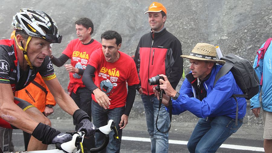 Lance Armstrong, con casco blanco y maillot negro, compite en bicicleta. Varios espectadores, algunos con camisetas rojas con la palabra "ESPAÑA" y el año "2010", observan la carrera.