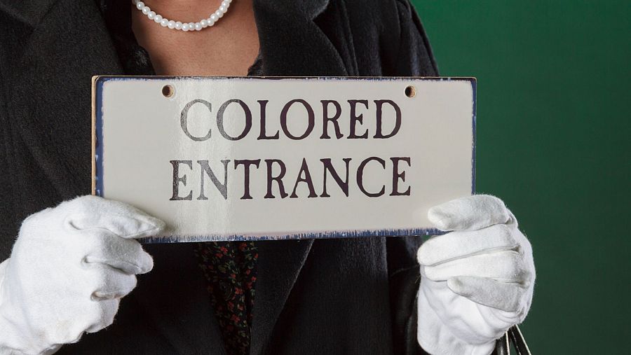 Una persona con guantes blancos sostiene un letrero con la inscripción "COLORED ENTRANCE". El letrero, con borde azul, parece antiguo y la persona lleva un abrigo negro y una blusa con estampado floral.