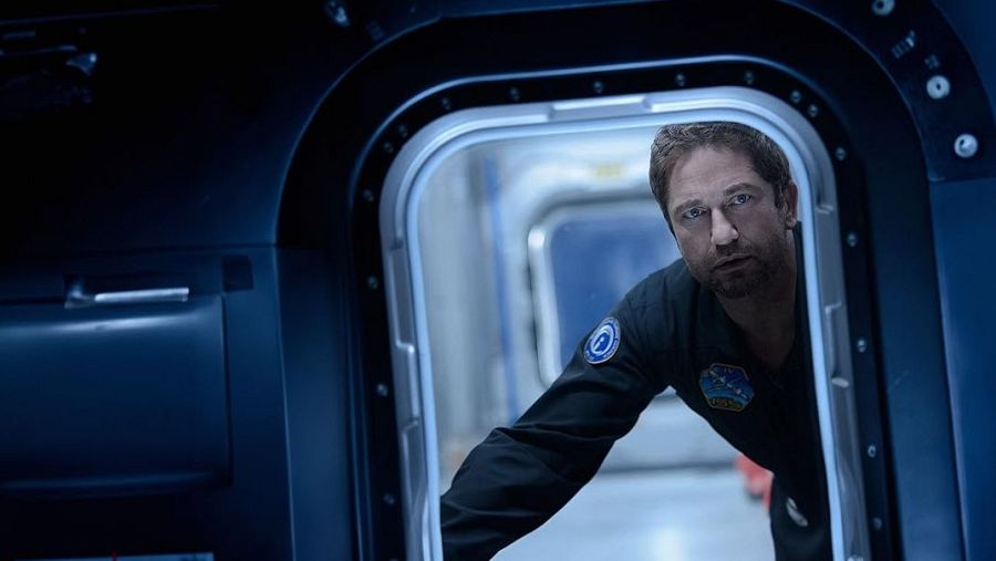 Un astronauta con barba, ataviado con un traje espacial oscuro, mira fijamente a través de una escotilla, con el interior de la nave espacial visible en tonos fríos.