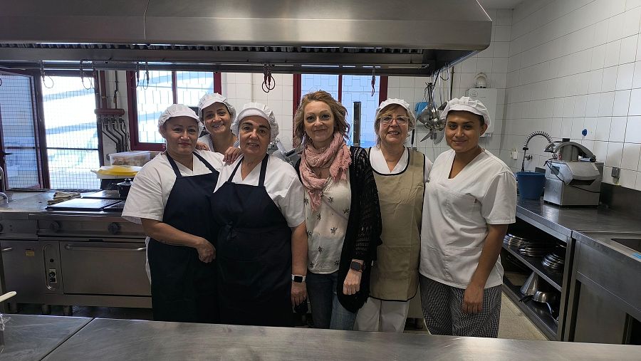 En una cocina con paredes blancas y electrodomésticos de acero inoxidable, un grupo de mujeres posa. Cinco de ellas visten atuendos de cocina, mientras que una mujer en el centro se distingue por su vestimenta.