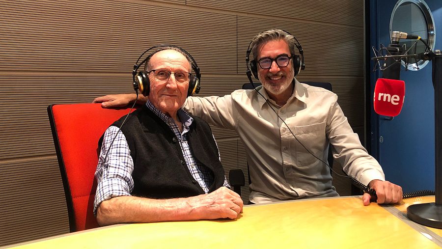Dos hombres sonríen en un estudio de radio. Uno de ellos, mayor, está sentado con auriculares y camisa de cuadros, mientras que el otro, más joven, lo abraza. Frente a ellos, una mesa amarilla y un micrófono con el logo de una emisora.