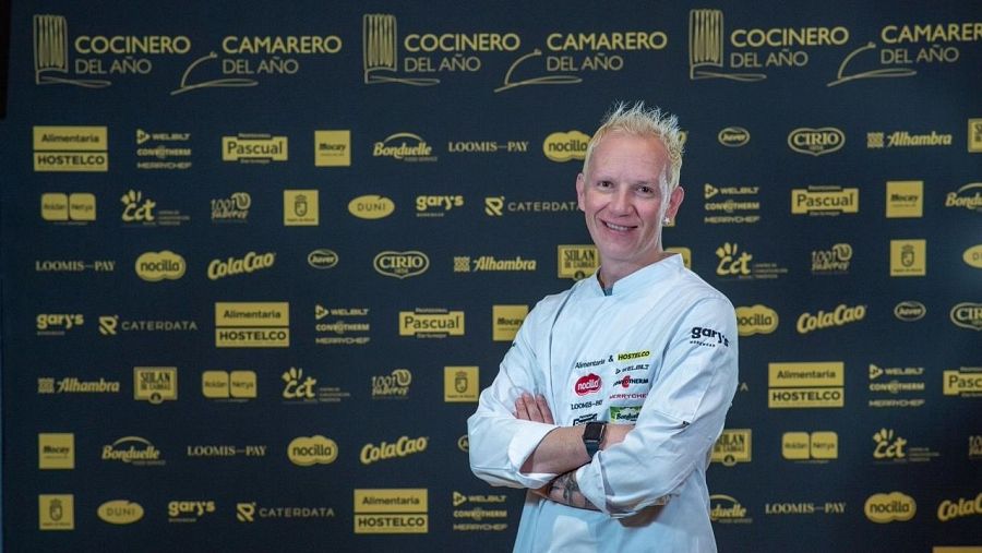 Toño Rodríguez, con chaqueta de chef y cabello rubio platino, sonríe a la cámara. El fondo negro muestra logos de marcas y los textos "COCINERO DEL AÑO" y "CAMARERO DEL AÑO".