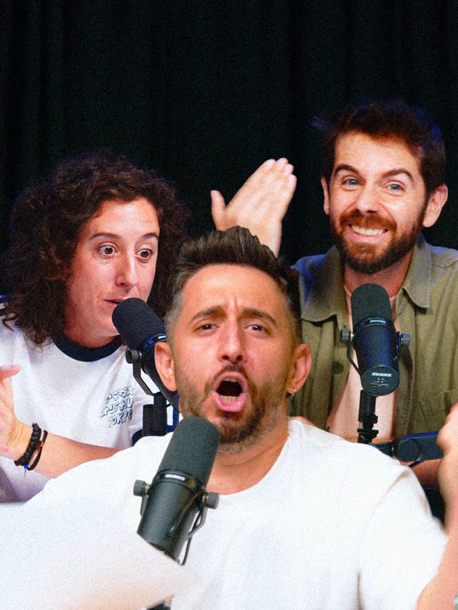 Tres hombres en un estudio de grabación, posiblemente un podcast, interactúan. Uno de ellos, con barba y camiseta blanca, habla y gesticula, mientras que los otros dos, uno con pelo rizado y otro con barba, aplauden y sonríen.