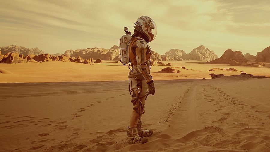 En un desierto de arena rojiza, un astronauta con un traje espacial blanco y naranja, y una mochila con el número "2", contempla el horizonte. El cielo está nublado y se aprecian huellas en la arena.