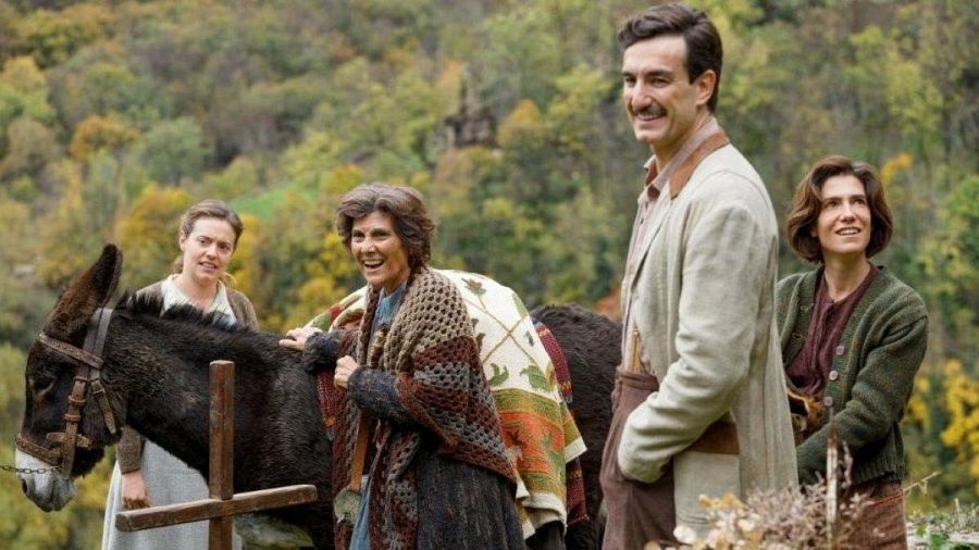 Un grupo de cuatro personas y un burro se encuentran en un entorno montañoso. Una mujer joven con vestido claro y chaleco, junto a un burro, está acompañada por una mujer mayor con chal floral, un hombre con bigote y chaqueta crema, y una mujer con cárdigan verde.