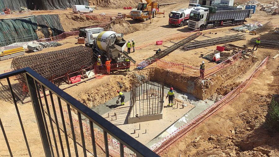 En la imagen se aprecia una obra en construcción en el lecho de un barranco, con una estructura de hormigón en proceso. Se distinguen trabajadores, varillas de acero, una hormigonera y maquinaria pesada.