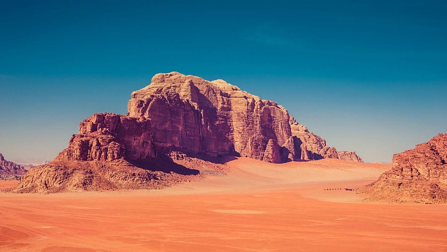 Un paisaje desértico con montañas rocosas rojizas bajo un cielo azul claro, con sombras suaves y una superficie arenosa ondulada por el viento.