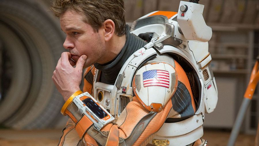 Matt Damon, con un traje espacial, se toca la barbilla con una expresión pensativa. En el hombro del traje se aprecia la bandera de Estados Unidos.